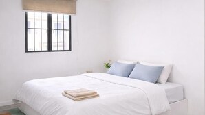 Ein Schlafzimmer mit Bett, einem Fenster mit Jalousien, einem Nachttisch mit Pflanze und einem hellen Boden.