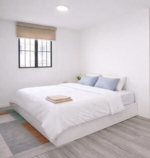 Une chambre à coucher avec un lit, une fenêtre avec des stores, une table de chevet ornée d’une plante et un plancher de couleur claire.