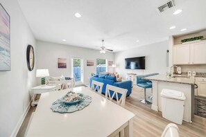 A modern kitchen with a TV, a dining area with a table and chairs, a blue sofa, and a ceiling fan. - Rae of Sunshine (Panama City Beach)