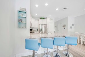 A modern kitchen with a granite countertop, four blue bar stools, white cabinetry, and a stainless steel refrigerator. - Rae of Sunshine (Panama City Beach)