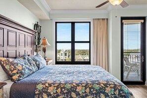 A bedroom with a large bed, a nightstand, a lamp, a ceiling fan, a window with a view of houses, and a door leading to a balcony. - Enjoy the best of Destin at the Harbor Lookout by Rockstar Retreats! (Destin)