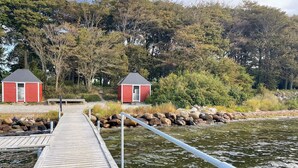 Une jetée en bois mène à deux cabanes rouges aux toits gris, entourées d’une forêt dense et bordées par une étendue d’eau calme.