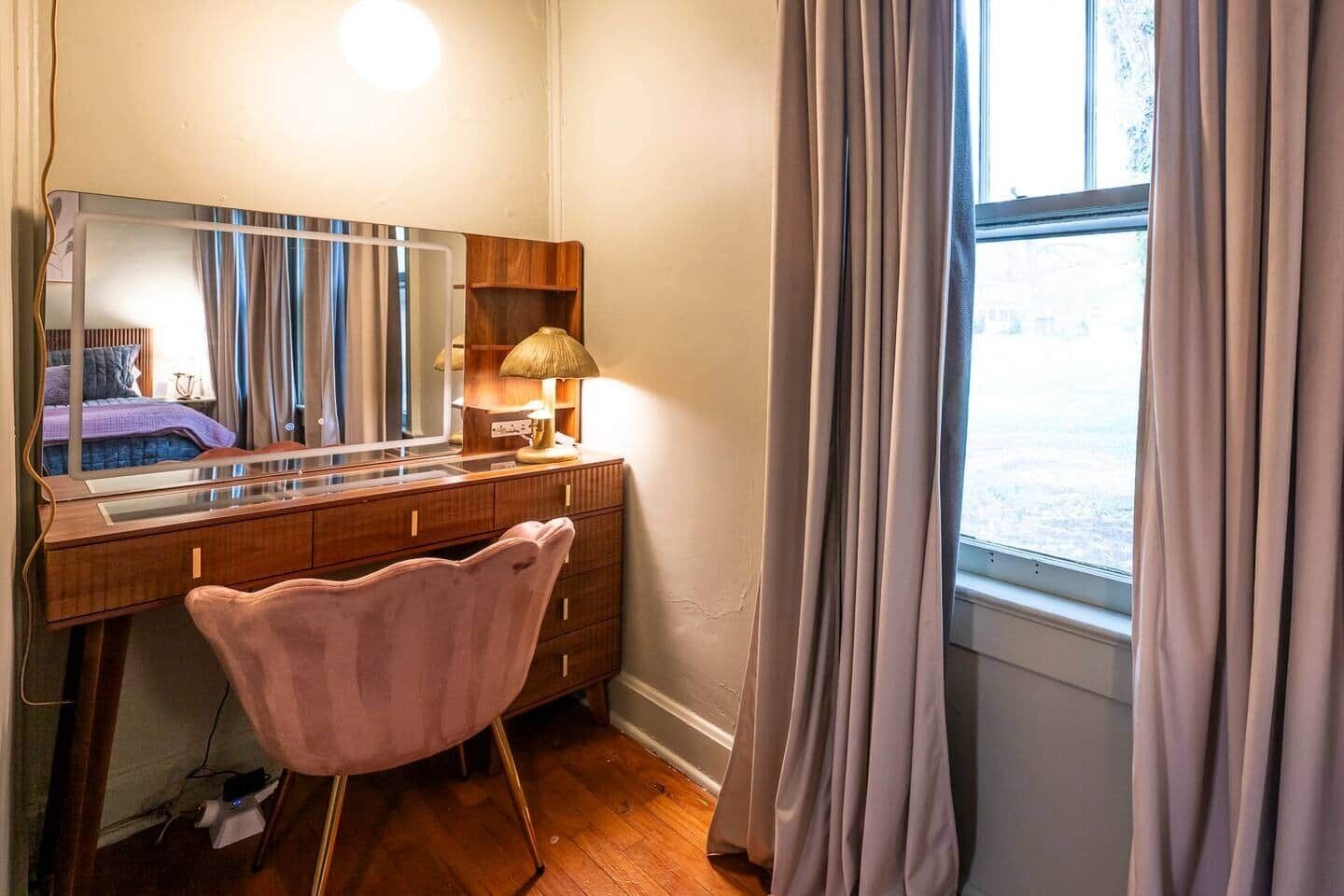 Un dormitorio con un aparador de madera, un tocador con espejo, una silla rosa y una ventana con cortinas.