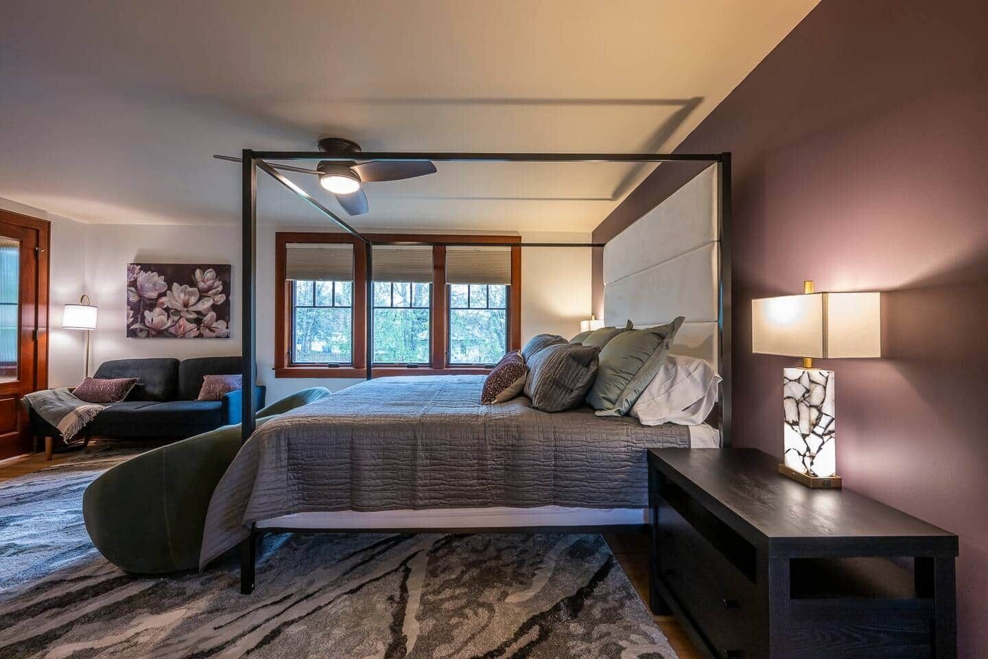 A bedroom with a four-poster bed, a ceiling fan, a large window, and a nightstand with a lamp.