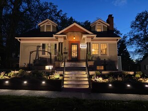 A two-story house with a front porch, a staircase, and a well-lit entrance. - Luxe Historic Gem: Hidden Game Rm, Hot Tub, Sauna (Lee's Summit)