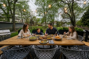 A wooden dining table with chairs, surrounded by greenery and trees, with a grill and outdoor lighting.