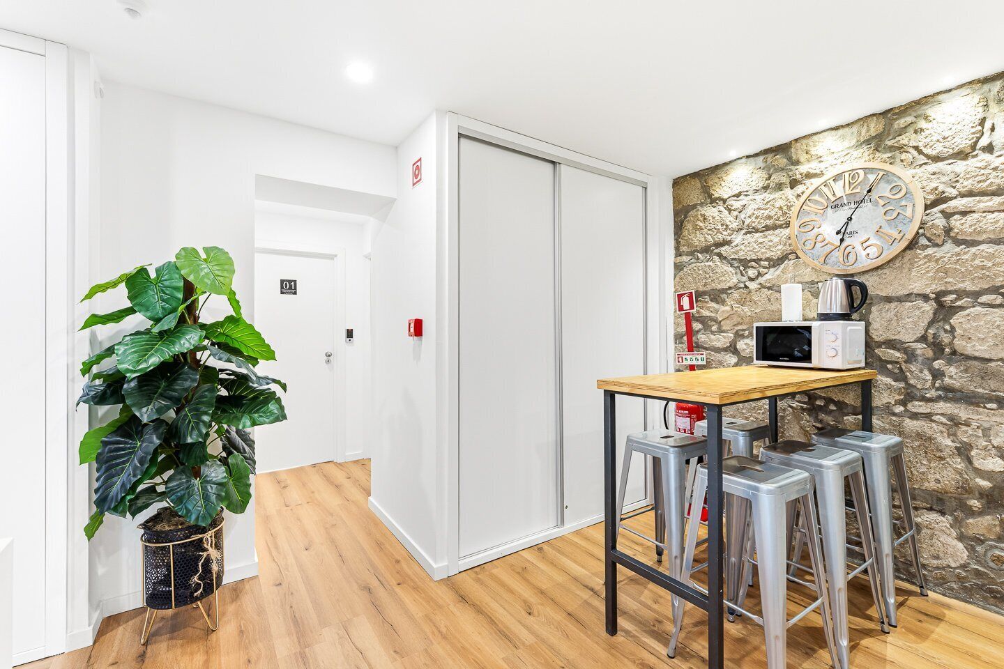 A hotel room with a wooden floor, a table with a microwave, a clock on the wall, and a potted plant.
