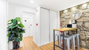 A hotel room with a wooden floor, a table with a microwave, a clock on the wall, and a potted plant.