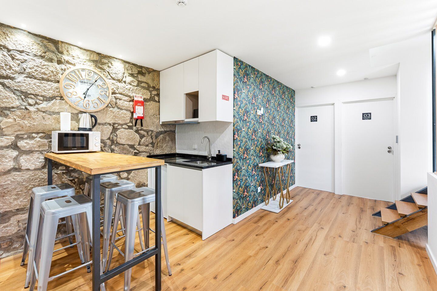 A modern kitchen with a stone wall, a clock, a microwave, and a sink. There are stools and a small table.