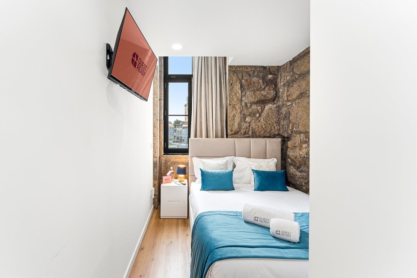 Een hotelkamer met een bed, nachtkastje, televisie, een accentmuur van steen en een raam met gordijnen.