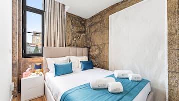 A hotel room with a stone wall, a bed with blue bedding, a nightstand with a lamp, and a window with curtains.