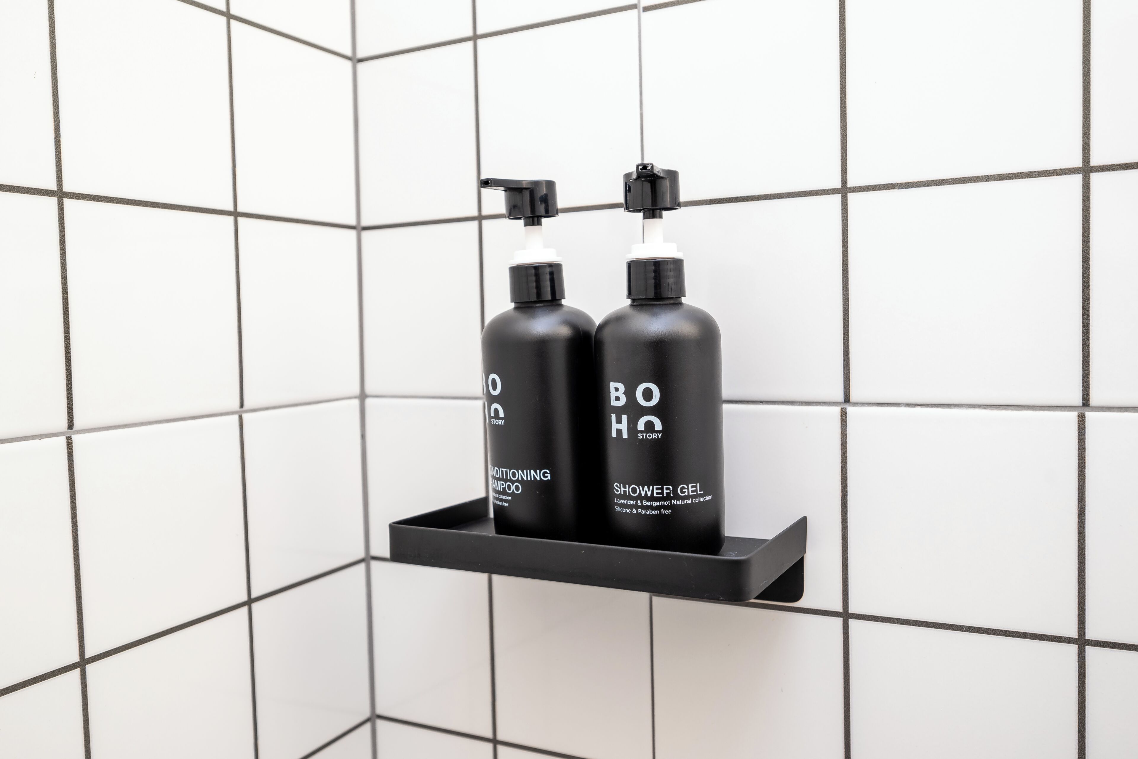 Deux flacons noirs de shampoing et de gel douche de la marque BOHO Story, posés sur une étagère noire devant un mur carrelé.