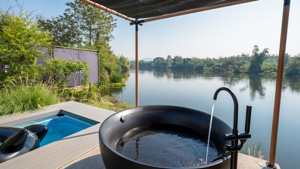 Ein Whirlpool mit Blick auf einen ruhigen See, umgeben von viel Grün und einer überdachten Terrasse.