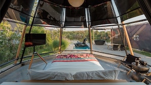 Eine Zelthütte mit Bett, Fernseher und Blick auf ein Gewässer und Grünflächen.