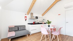 Een kleine keuken met een magnetron, fornuis en spoelbak, een grijze bank met een roze plaid, en een eethoek met roze stoelen en een tafeltje.