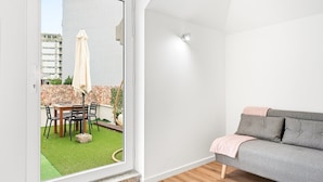 Een modern appartement met een schuifdeur naar een klein balkon met een tafel en stoelen, een grijze bank en een kleine tuin.
