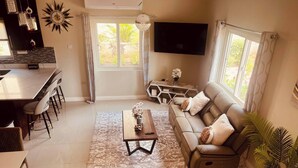 A modern living room with a sofa, coffee table, TV, and a dining area with chairs. - Tranquility Loft, Ocho Rios, Solar Powered (Ocho Rios)