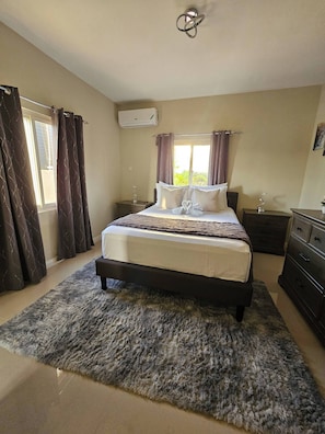 Une chambre à coucher comprenant un lit, une table de chevet, une commode, une fenêtre avec des rideaux et un appareil de climatisation.