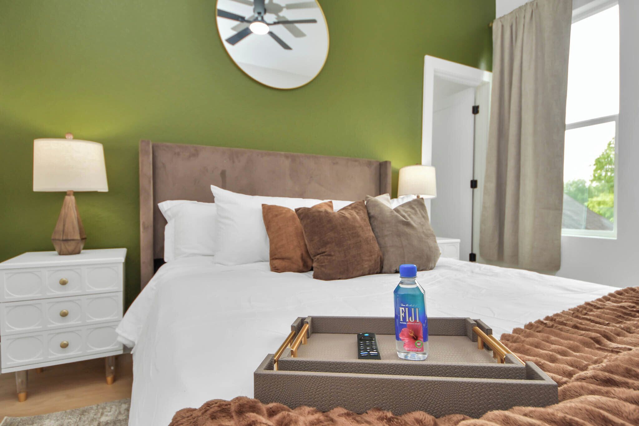Ein Hotelzimmer mit Bett, Nachttischen, einer Wanduhr, einem Fenster mit Vorhängen und einem Tablett mit Flasche und Fernbedienung.