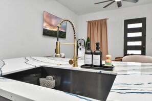 A modern kitchen with a gold faucet, marble countertop, and a sink. There are two bottles of soap and a wooden brush on the counter. - 2BR w Ensuite Baths + 7 Min to NRG (Houston)