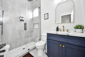 Ein modernes Badezimmer mit Duschkabine aus Glas, einer weißen Toilette, einem Waschtisch mit goldfarbenem Wasserhahn und einem Spiegel mit goldener Rahmung.