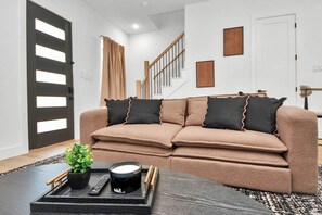 A living room with a brown sofa, a black tray with a remote, a plant, and a coffee cup. - 2BR w Ensuite Baths + 7 Min to NRG (Houston)