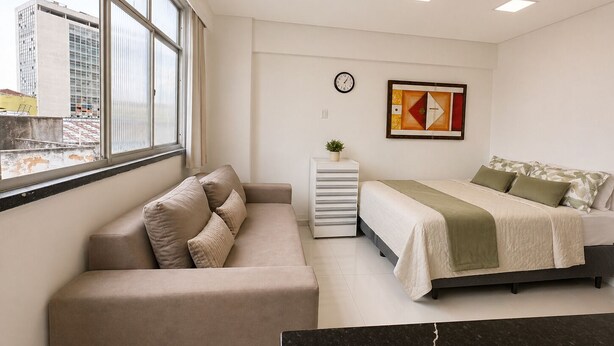 A modern bedroom with a bed, a sofa, a small side table, and a window with a city view. - Studio apartment in the city center with Wi-Fi. (Manaus)