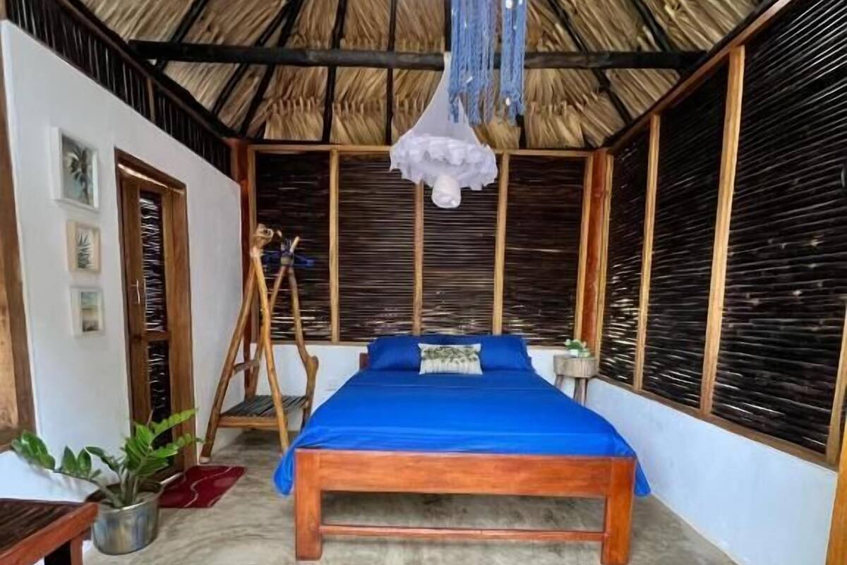 Ein Zimmer mit einem Bett, einer blauen Matte mit der Aufschrift 'BIENVENIDO', einer Hängelampe und einem Strohdach.