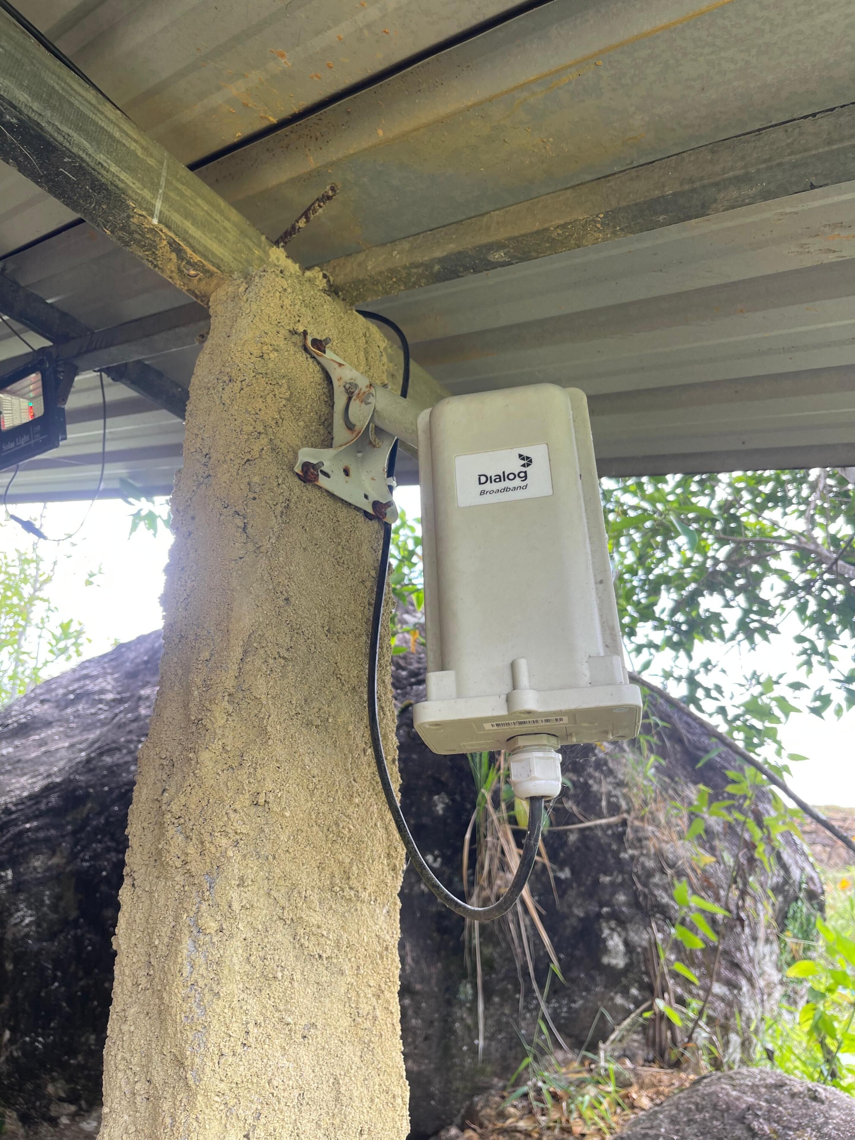 Una caja de servicios identificada con 'Dialog Broadband' adherida a un pilar de hormigón bajo un techo.