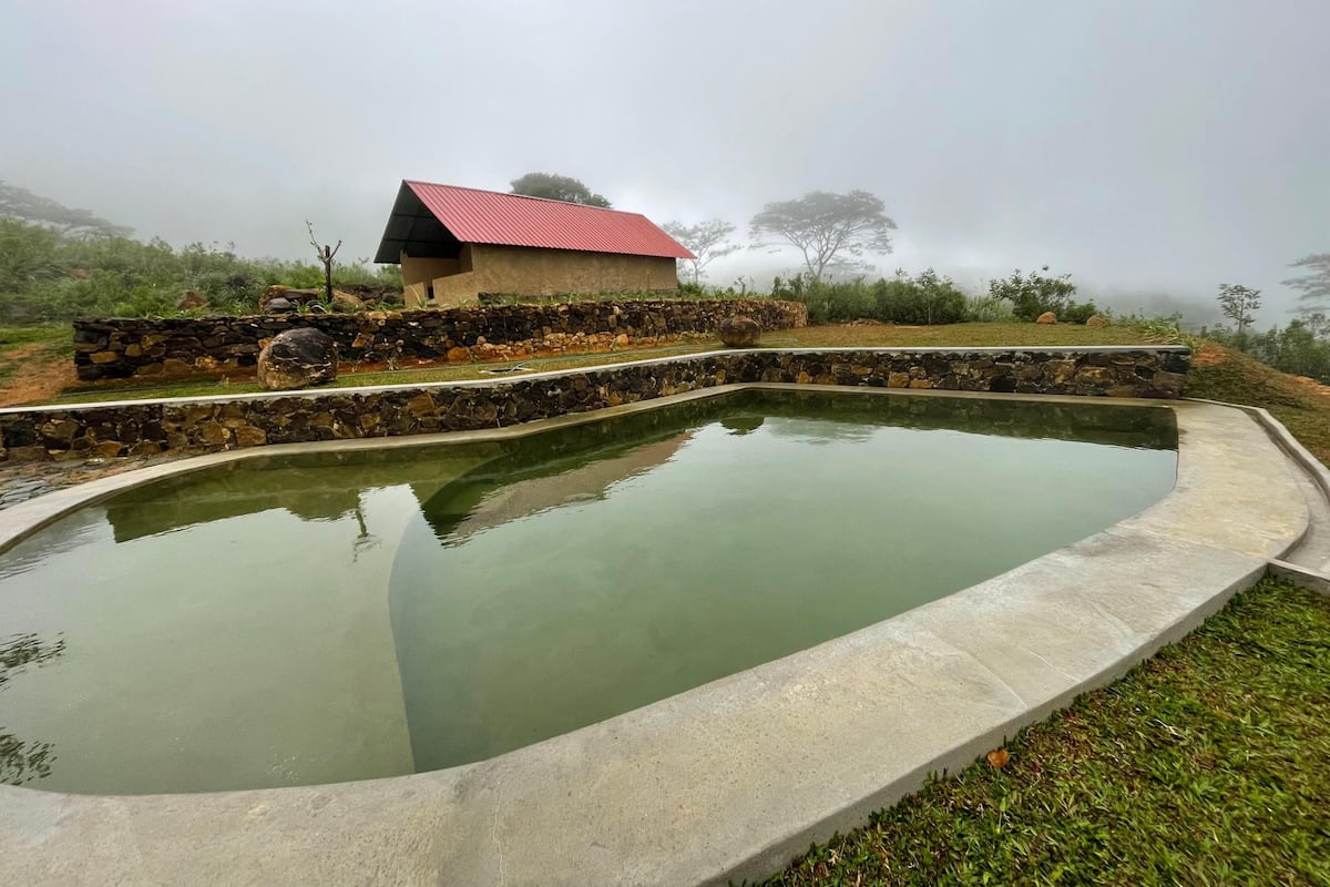 Una piscina con borde de piedra, un pequeño edificio con techo rojo y un muro de piedra en un paisaje brumoso.