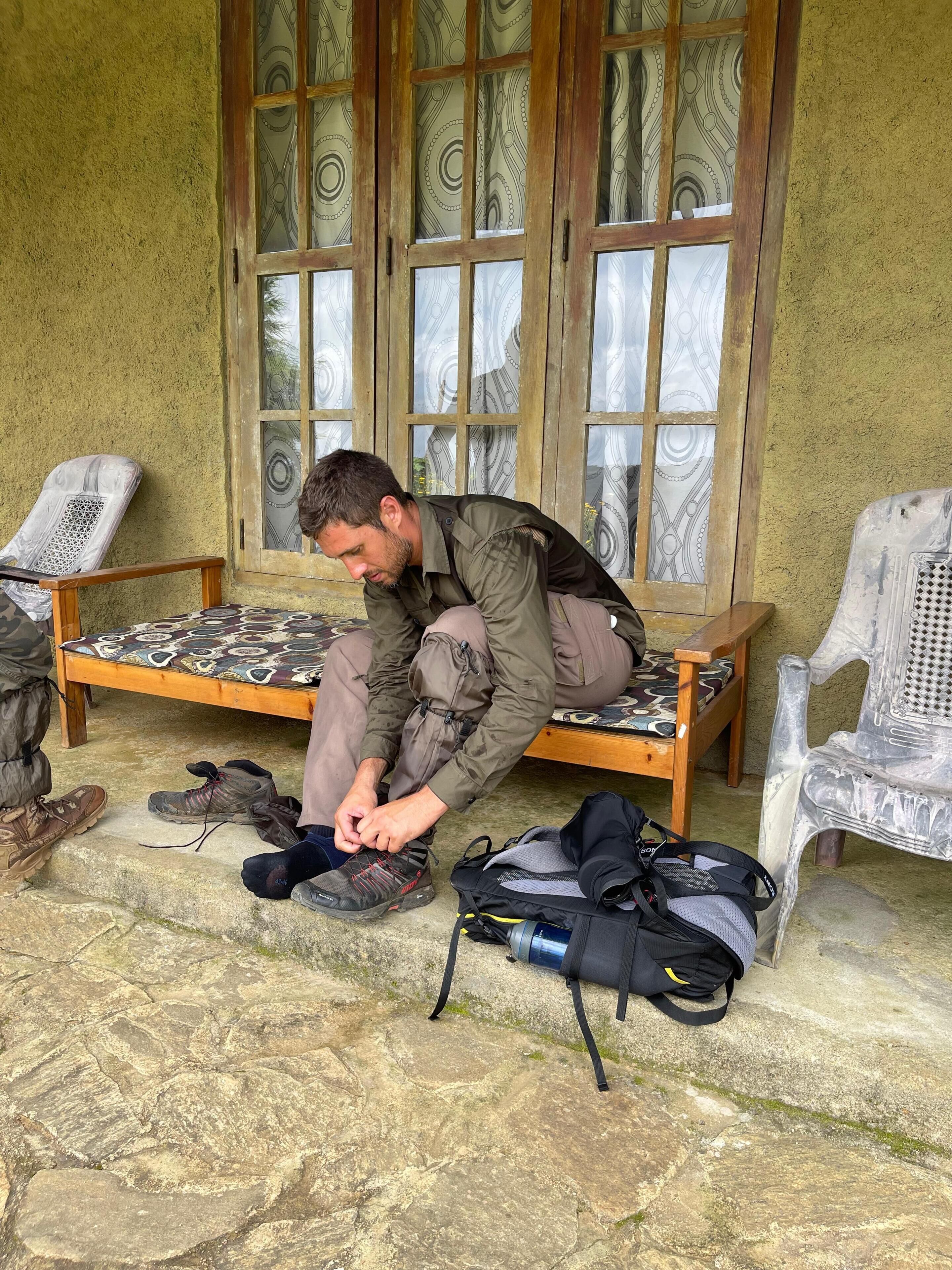 A person sitting on a bench, tying their shoe, with a backpack and another pair of shoes nearby.