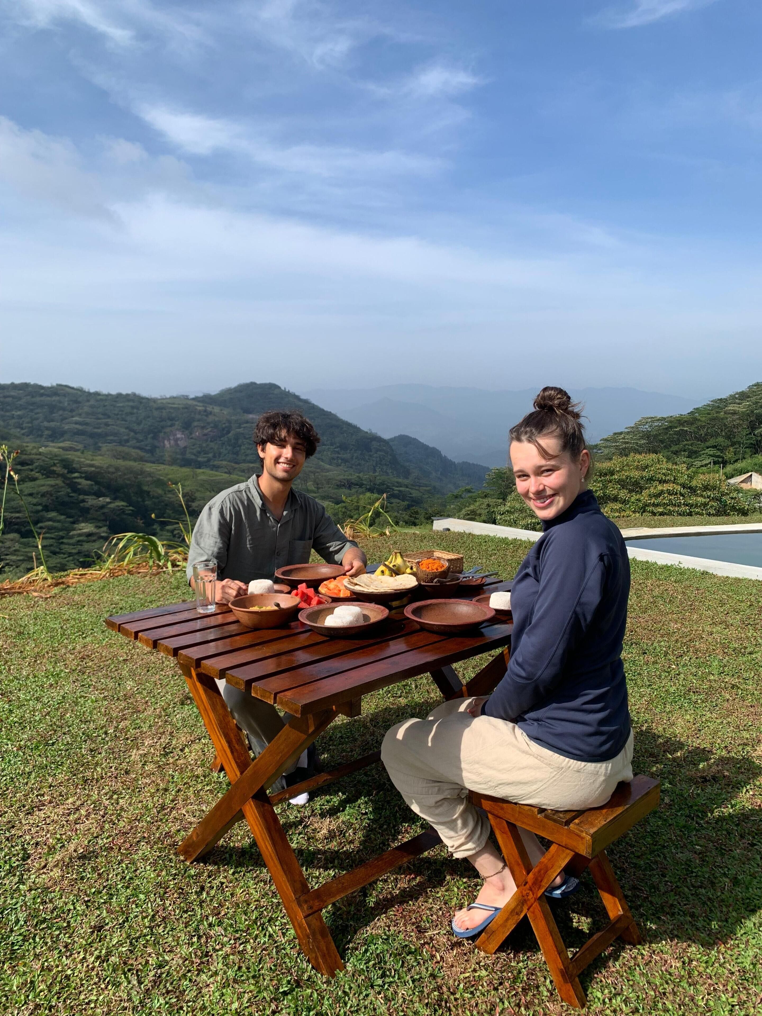 Two people dining at an outdoor table with a scenic view of hills and a road in the background.
