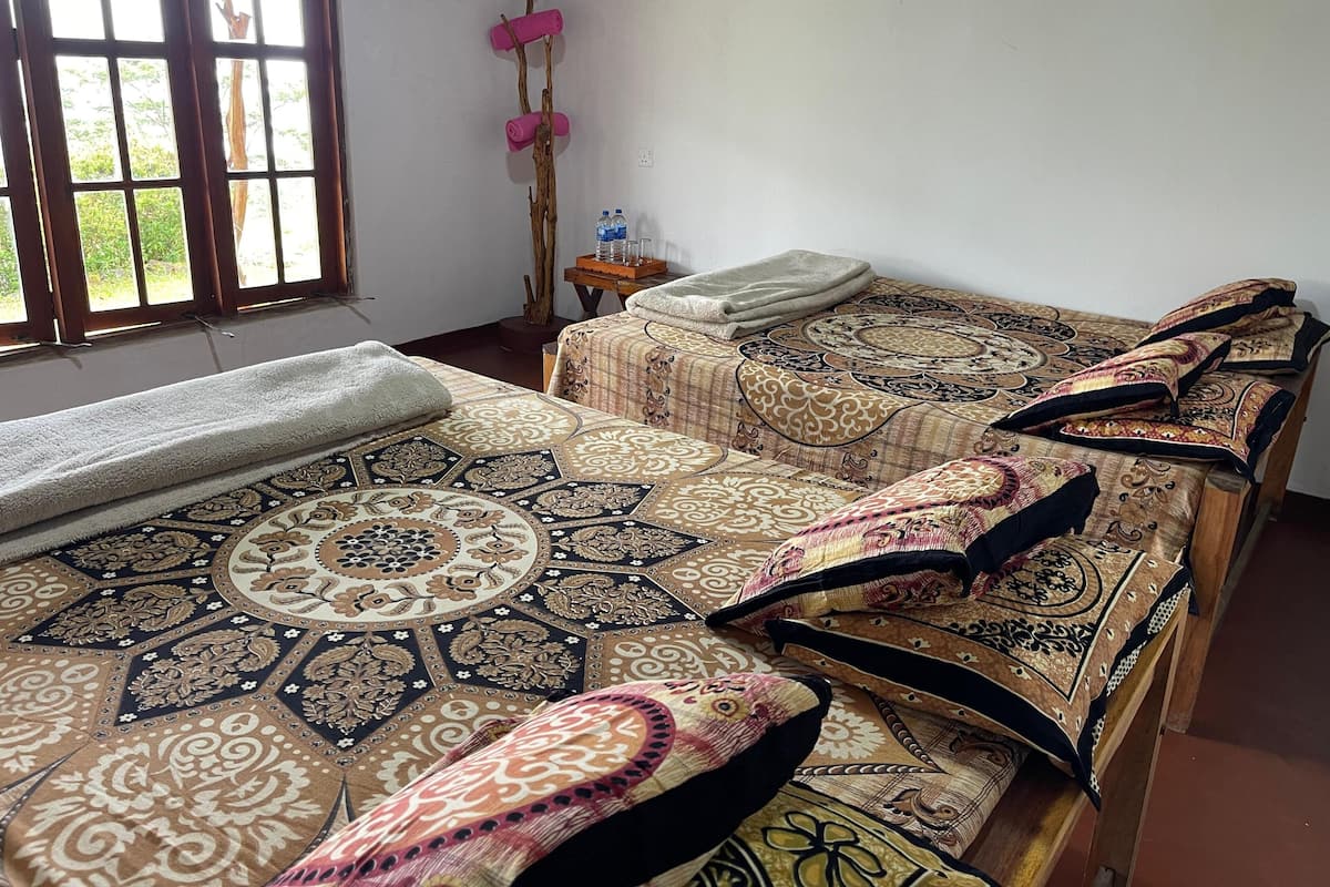 Una habitación con dos camas, una ventana con cortinas, una mesita con una botella de agua y un perchero en la pared.