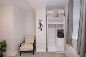 Ein Zimmer mit einem Sessel, einem weißen Schrank mit Regalen, einer hängenden Pflanze und einem Fenster mit Vorhängen.