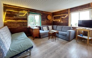 A rustic cabin interior with a sofa, a small table, a television, and a window with a view of greenery.