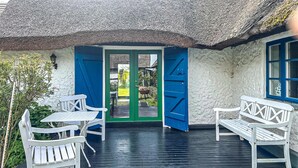 Ein reetgedecktes Häuschen mit einer Holterrasse, weißen Möbeln und einer grünen Tür.