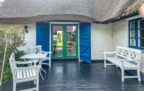 Un chalet au toit de chaume, doté d’une terrasse en bois, de meubles blancs et d’une porte verte.