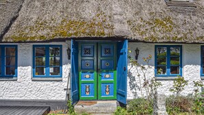 Ein traditionelles reetgedecktes Häuschen mit weißer Fassade und blauen Akzenten, mit einer zentralen Tür mit dekorativen Verzierungen und zwei Fenster mit blauen Rahmen.