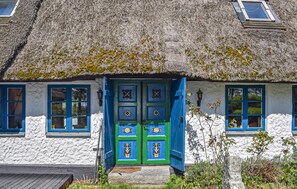 Une maison de campagne traditionnelle à toit de chaume, aux murs blancs et aux détails bleus, avec une porte centrale ornée de panneaux décoratifs et deux fenêtres aux cadres bleus.