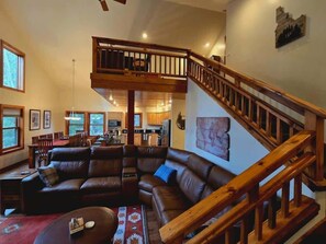 Ein geräumiges Wohnzimmer mit einer großen, im L-förmigen Verlauf angeordneten Couch, einer Holztreppe und einer Küche im Hintergrund.