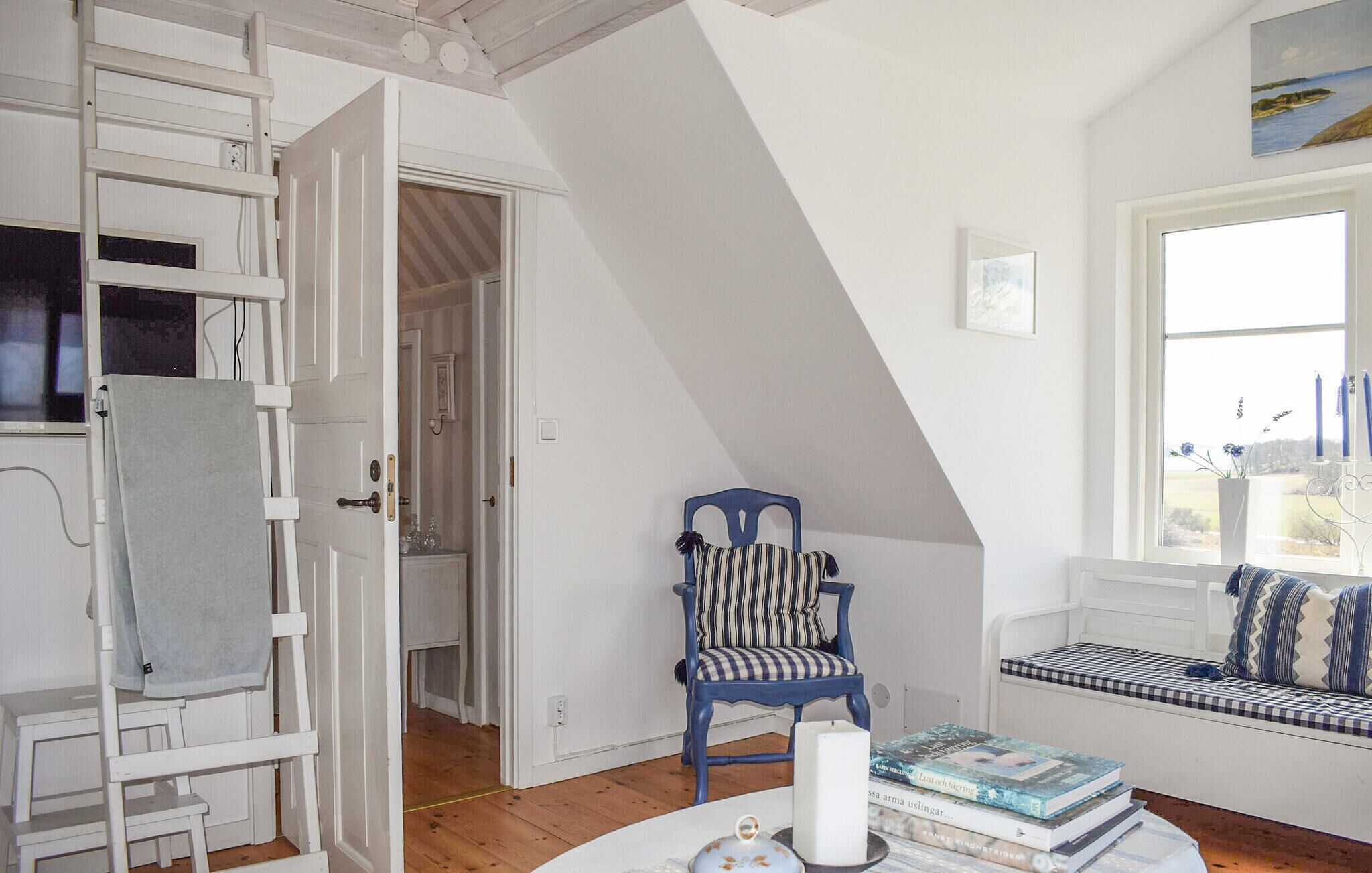A room with a lofted sleeping area, a chair with striped cushions, a bedside table with books, and a window with a view.