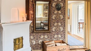 Une pièce avec un miroir, une cheminée, un coffre en bois et une porte donnant sur une autre pièce.