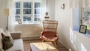 Una sala de estar acogedora con un sofá, una mesa de centro de madera, un sillón de mimbre y un ventanal con vistas a árboles.