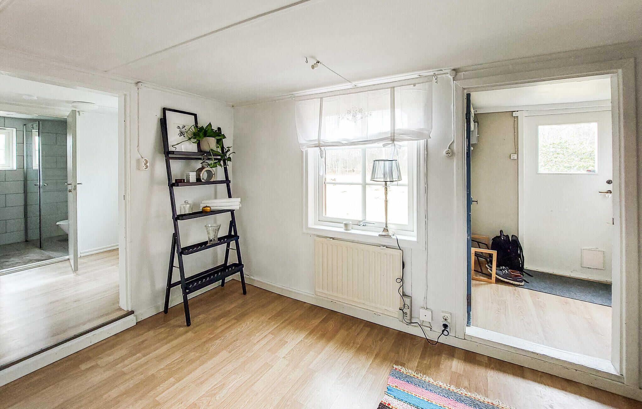 A room with a wooden floor, a radiator, a window with curtains, a shelf with items, and a door leading to another room.