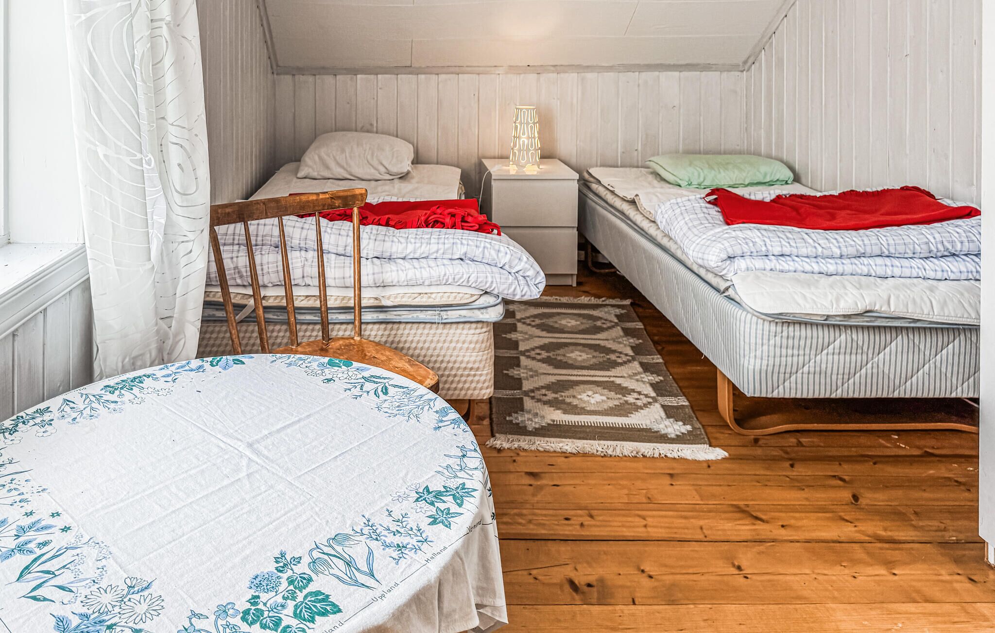 A small, cozy room with two beds, a wooden chair, a table, and a nightstand with a lamp.