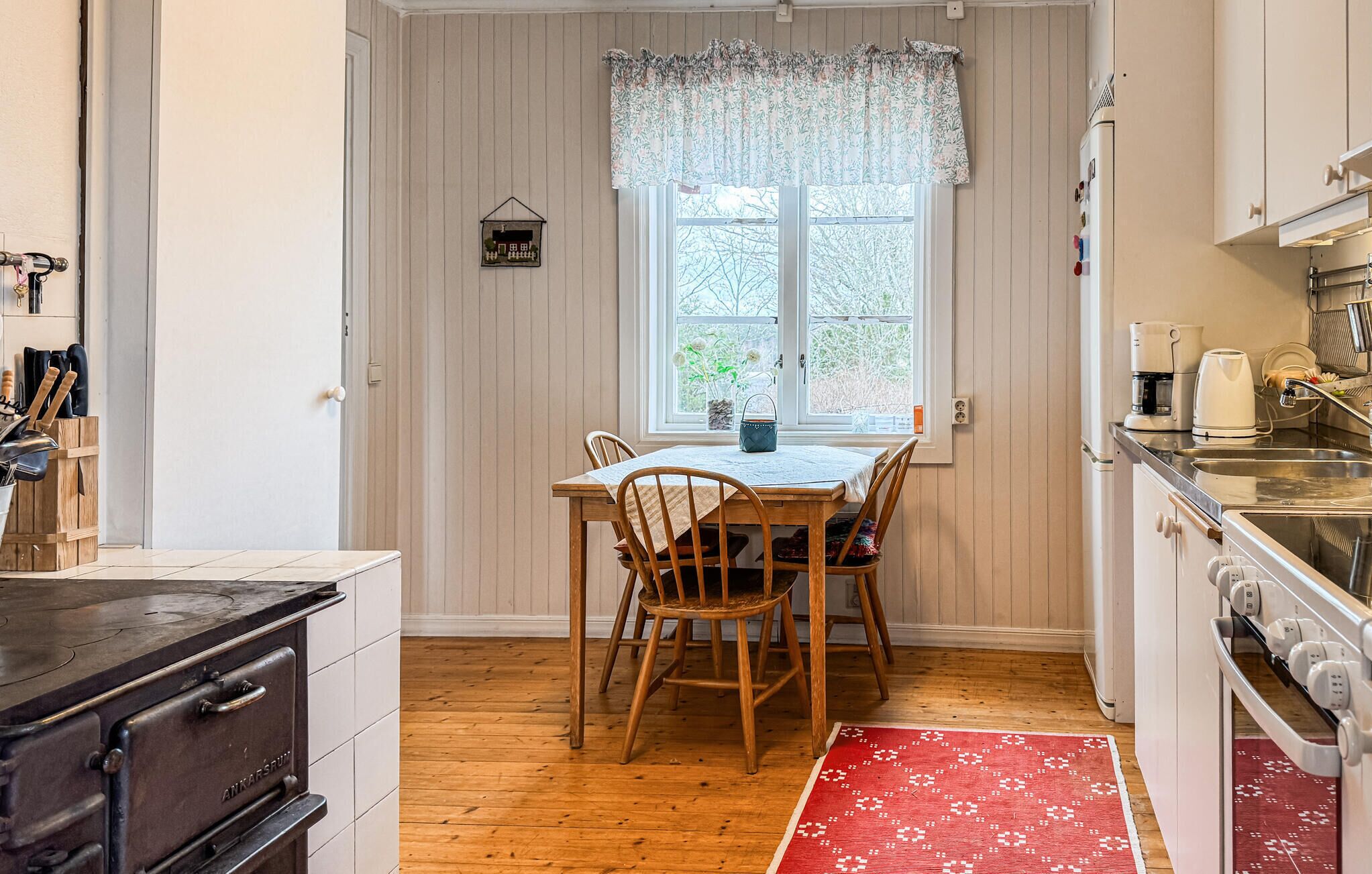 A kitchen with wooden cabinets, a stove, a window with a floral curtain, and a dining table with chairs.