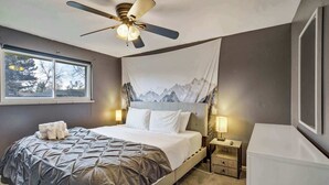 Um quarto com uma cama, um ventilador de teto, uma janela, uma mesinha de cabeceira com luminária e uma televisão presa na parede.