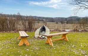 An outdoor seating area with a stone fireplace, two wooden benches, and a grassy field with trees in the background.
