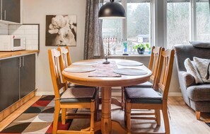 A dining area with a round table, six chairs, a black pendant lamp, a kitchen with a microwave, and a window with a view of trees and a house.