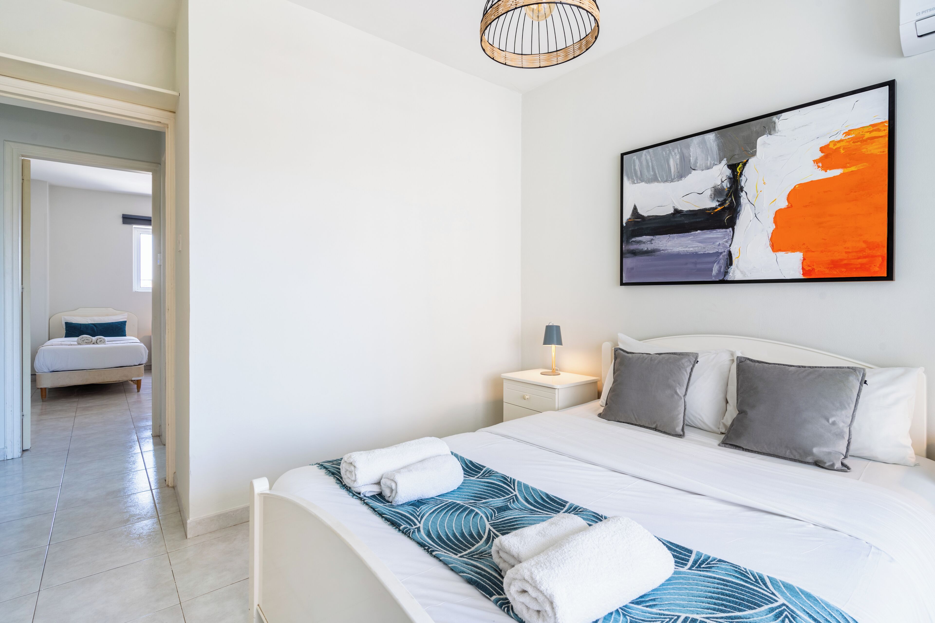 Un dormitorio moderno con una cama grande, una mesita de noche, un cuadro abstracto en la pared y un pasillo que lleva a otra habitación.
